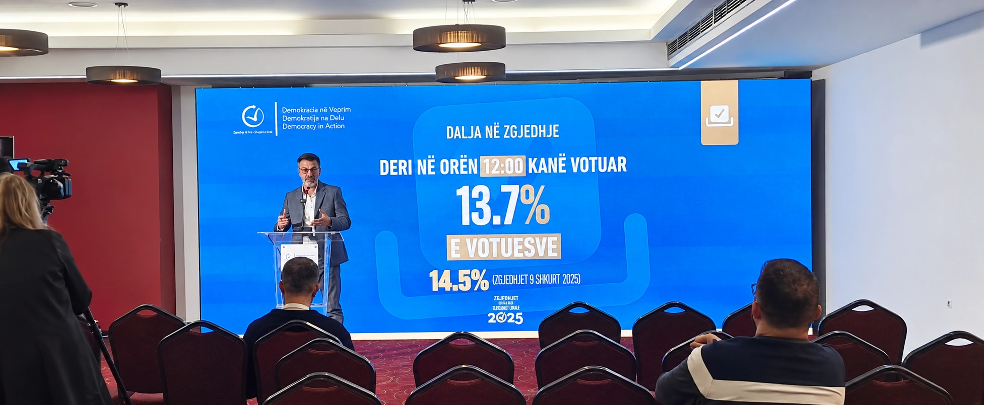 Conference speaker at a lectern in front of a large blue LED display